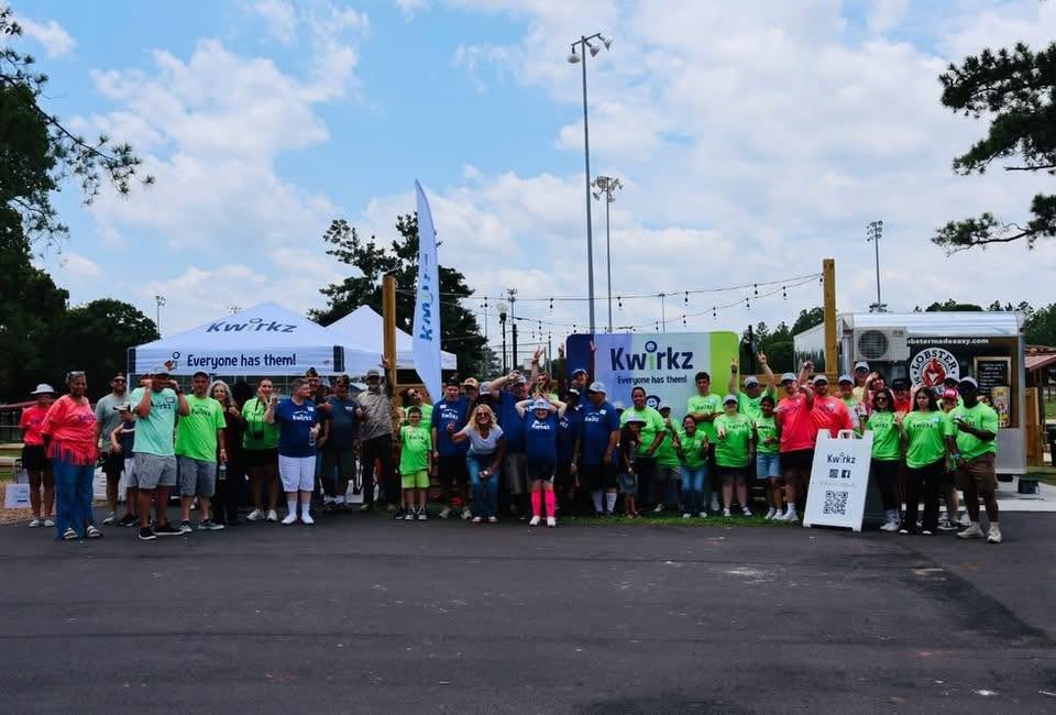 A crowd of roughly fifty Kwirkz participants, families and volunteers standing together outdoors with their arms raised, in front of a Kwirkz canopy and a banner reading Everyone has them!, with a food truck and string lights behind them.