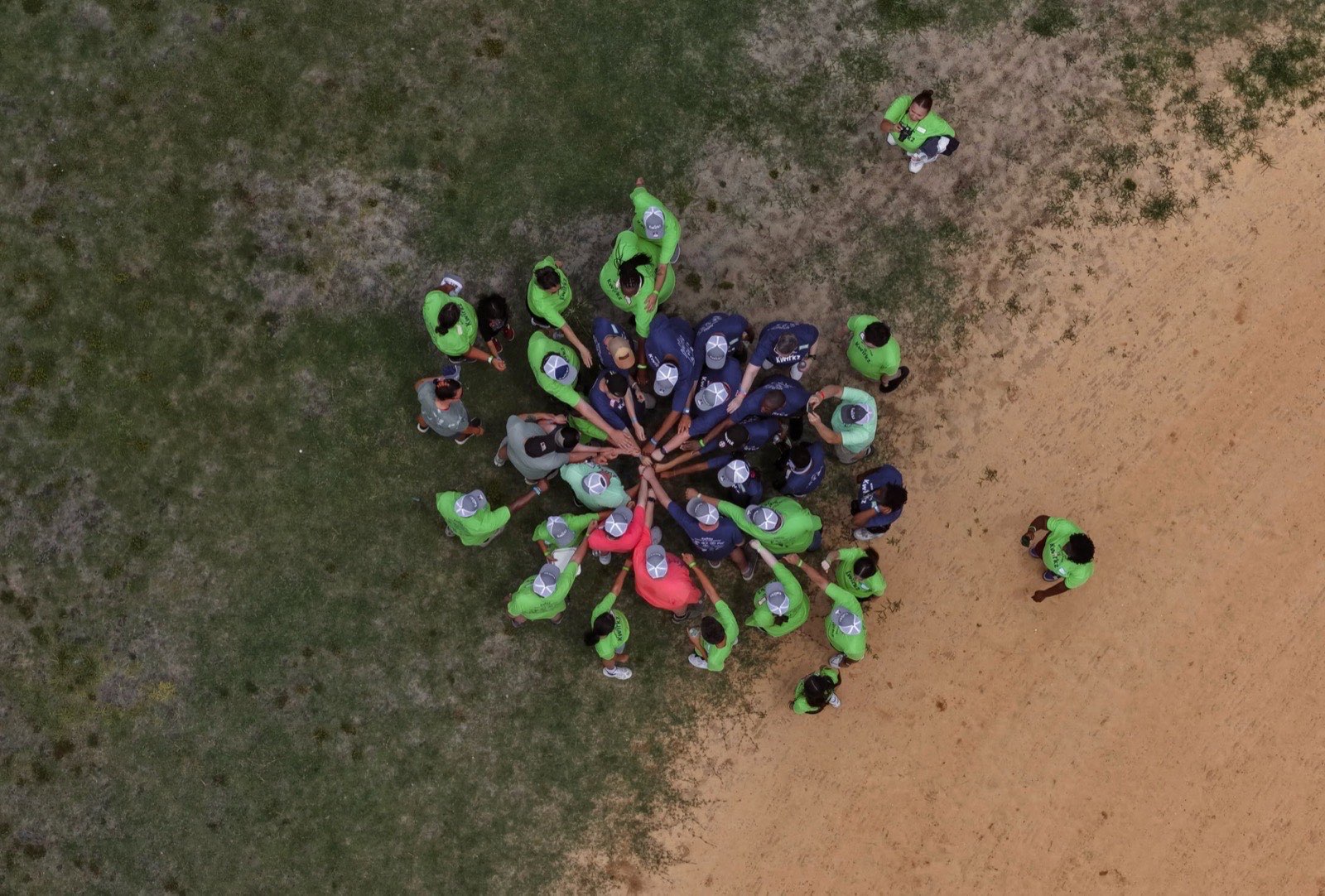 Overhead view of about thirty Kwirkz participants and volunteers standing in a circle on a grass field with their hands stacked together in the middle. Participants wear navy Kwirkz shirts, volunteers wear bright green ones, and nearly everyone is wearing a grey Kwirkz cap.