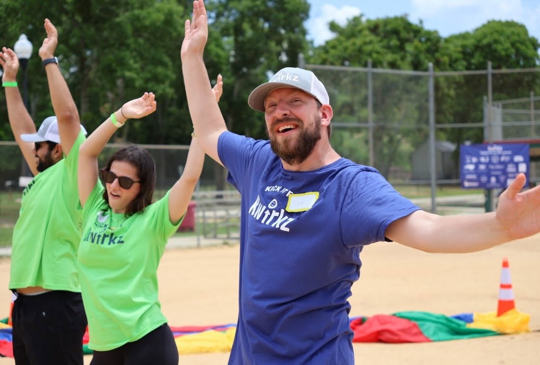 A bearded man in a navy Kick it for Kwirkz shirt and grey Kwirkz cap with both arms thrown wide, mid-cheer, with two volunteers in bright green Kwirkz shirts celebrating behind him and a rainbow parachute laid out on the sand.