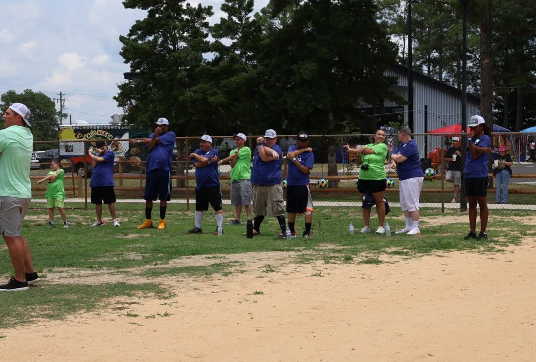 A line of about a dozen participants in navy Kwirkz shirts and grey caps doing arm-across-chest stretches on the grass, with a taco food truck and vendor tents visible behind the fence.