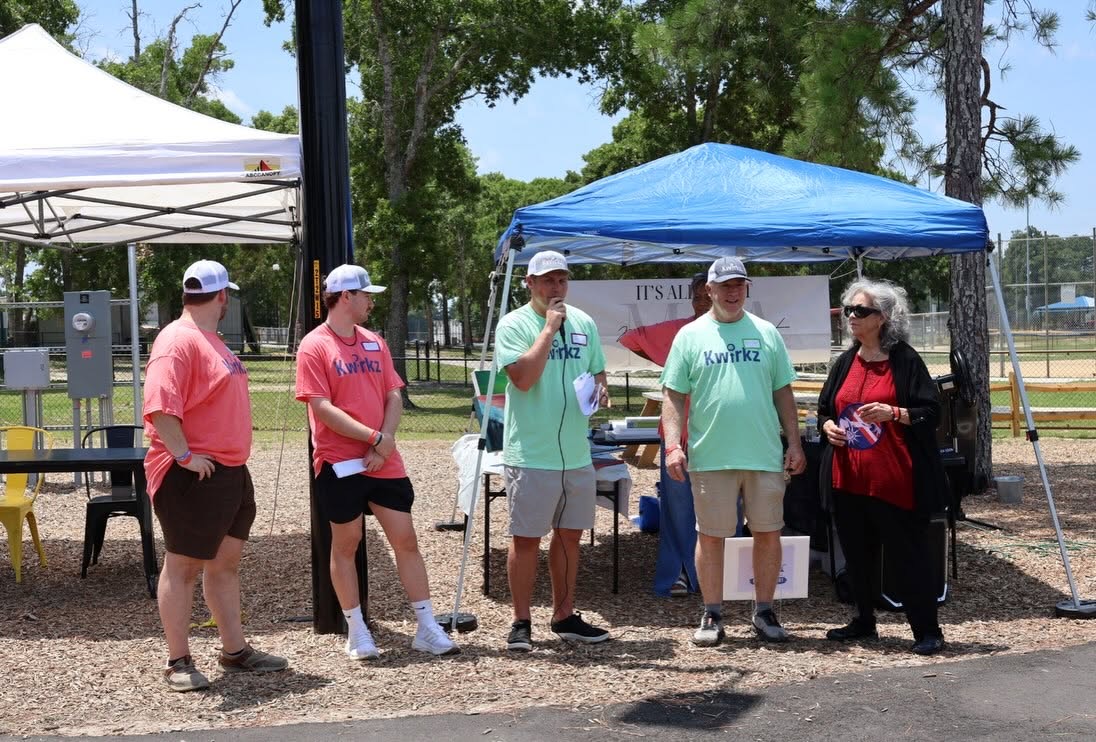 Four people in coral and mint Kwirkz shirts standing under pop-up canopies, one speaking into a handheld microphone, with an older woman in red standing beside them.
