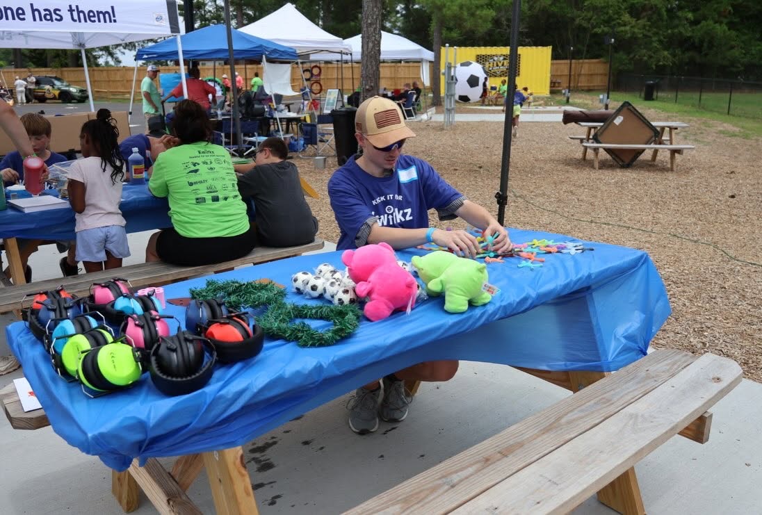 A volunteer in a navy Kick it for Kwirkz shirt setting out sensory supplies on a picnic table covered in a blue cloth: about a dozen pairs of noise-reducing ear covers in bright colours along the front edge, plus fidget toys and soft toys. The Hive building and a giant soccer ball are visible behind him.