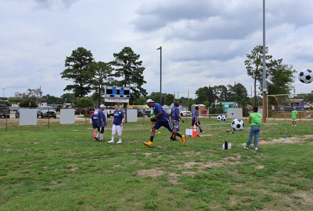 Participants in navy Kwirkz shirts playing soccer on a grass field, one mid-stride toward the ball, with oversized soccer balls, a volunteer in a green shirt watching, and a scoreboard behind them.