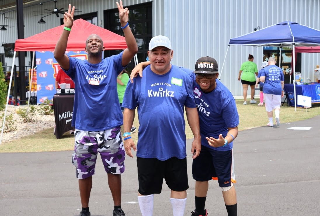 Three participants in navy Kick it for Kwirkz shirts posing together on the tarmac, arms raised and grinning, with vendor canopies and tables behind them.