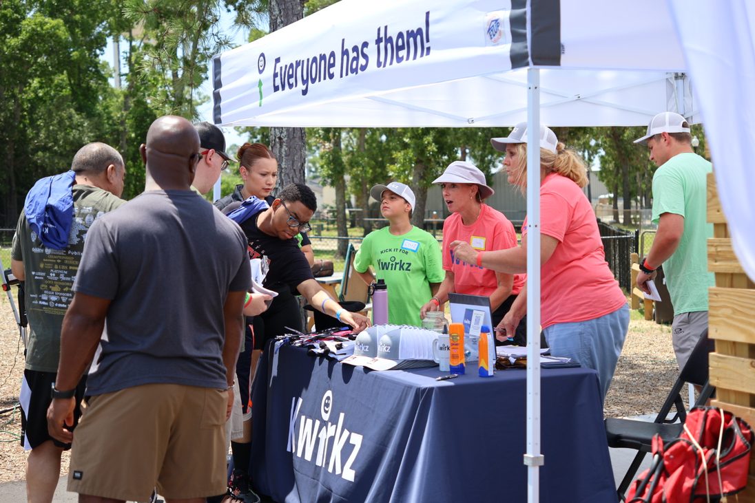 Volunteers working the Kwirkz welcome tent at Kick it for Kwirkz. A young volunteer in a green Kick it for Kwirkz tee and two volunteers in coral shirts hand out sunglasses and water to a queue of families across a table covered in a navy Kwirkz cloth. The canopy banner above reads Everyone has them.