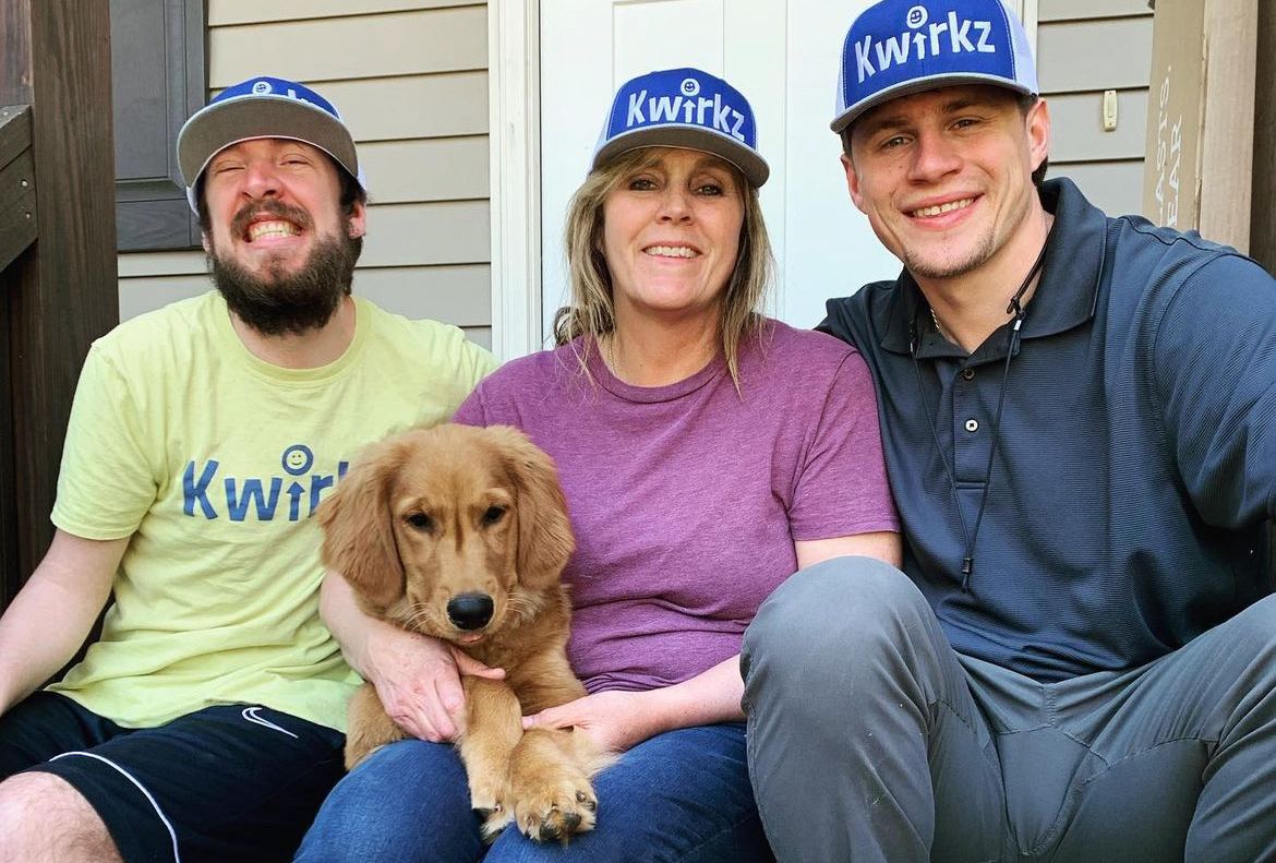 Three people sitting close together on a porch step, all wearing blue Kwirkz caps, with a golden retriever puppy resting across their laps. The person on the left wears a yellow Kwirkz t-shirt.